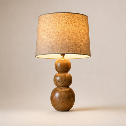 A table lamp with a three-tiered wooden base and a textured beige linen drum shade, shown lit against a neutral beige background.
