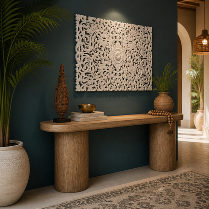 A styled interior scene with a carved white rectangular wall art piece mounted above a rustic wooden console table. The room has a deep teal wall, potted palm plants, decorative objects on the table, and a rug on the floor.