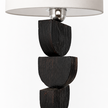 A modern table lamp with a sculptural base made of four stacked dark stained wooden blocks, topped with a brushed silver metal collar and a white drum shade, shown against a plain white background.